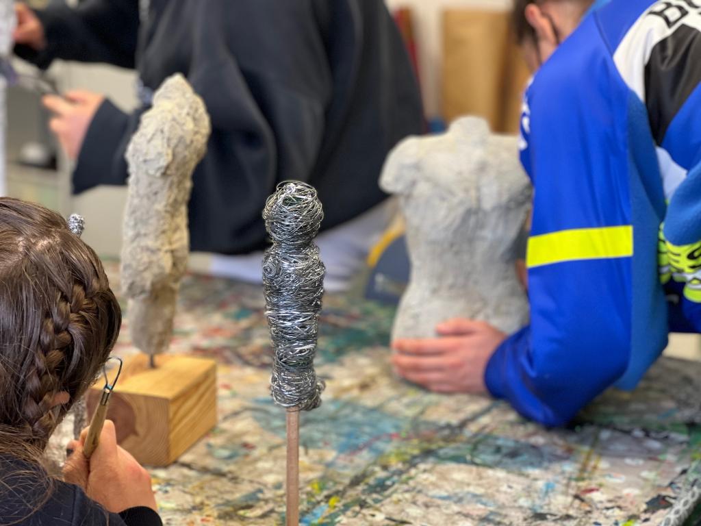 Zwei Personen arbeiten an einer Skulptur aus Stein. Einer von ihnen trägt ein blaues Hemd und hält die Skulptur mit beiden Händen. Auf dem Tisch liegen Werkzeuge und Materialien, die zur Bearbeitung der Skulptur verwendet werden.