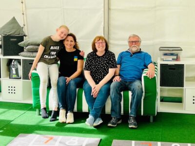 4 Personen sitzen auf einem grünen Sofa und lächeln in die Kamera. Das Sofa steht auf einem grünen Teppich und im Hintergrund sind weitere Möbel zu sehen.