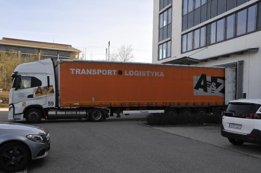 Ein orangefarbener Lastwagen mit der Aufschrift "Transport Logistik" steht auf einer Straße. Der Lastwagen ist groß und robust und bereit für den Transport von Waren. Im Hintergrund sind Gebäude und Bäume zu sehen.