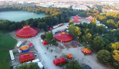 Luftaufnahme eines weitläufigen Parks mit mehreren roten Pavillons und üppigen grünen Bäumen. Die Anlage wirkt gepflegt und einladend. Im Hintergrund ist ein großer Fluss zu sehen, der die Landschaft durchzieht.
