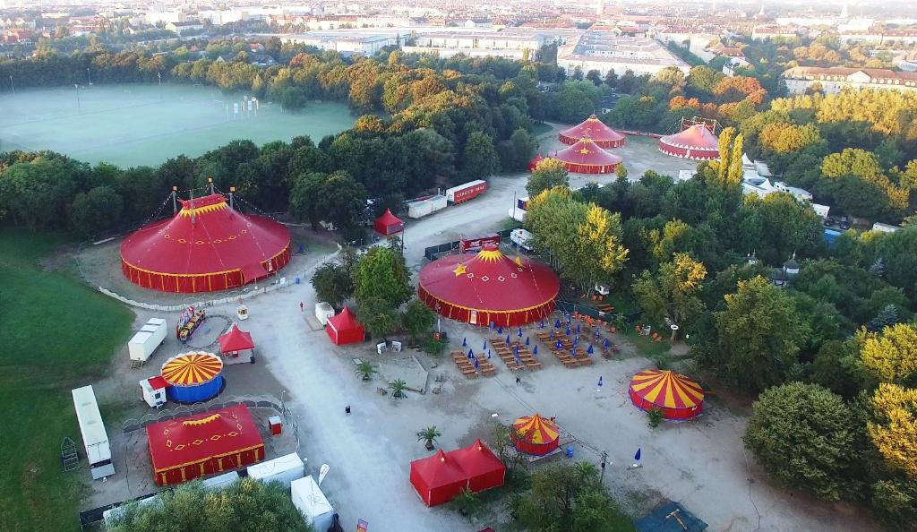Luftaufnahme eines weitläufigen Parks mit mehreren roten Pavillons und üppigen grünen Bäumen. Die Anlage wirkt gepflegt und einladend. Im Hintergrund ist ein großer Fluss zu sehen, der die Landschaft durchzieht.