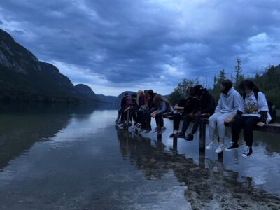 Eine Gruppe von Menschen sitzt auf einem Holzsteg, der in einen ruhigen See ragt. Der Himmel ist bewölkt und die umliegenden Berge sind im Hintergrund sichtbar. Die Szene wirkt ruhig und friedlich, während die Personen sich unterhalten und die Aussicht genießen.