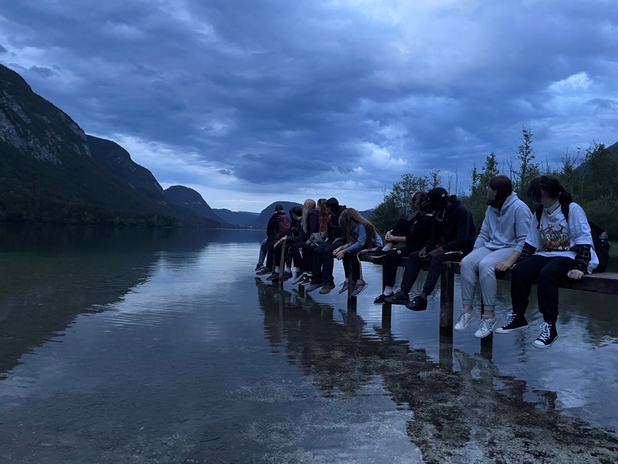 Eine Gruppe von Menschen sitzt auf einem Holzsteg, der in einen ruhigen See ragt. Der Himmel ist bewölkt und die umliegenden Berge sind im Hintergrund sichtbar. Die Szene wirkt ruhig und friedlich, während die Personen sich unterhalten und die Aussicht genießen.