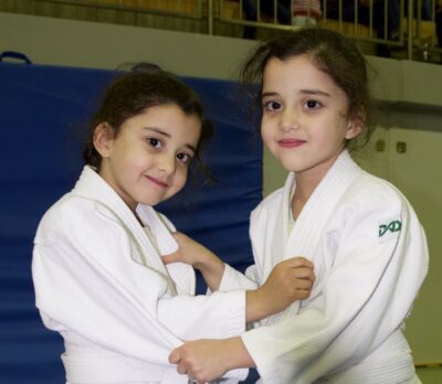Zwei Kinder in weißen Judoanzügen üben gemeinsam auf einer Matte. Sie halten sich gegenseitig fest und konzentrieren sich auf ihre Techniken. Im Hintergrund sind eine blaue Matte und eine Tribüne zu sehen.