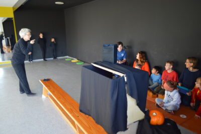 Eine Gruppe von Kindern sitzt auf dem Boden und auf einer Bank, während sie einer Person zuschauen, die eine Art Präsentation oder Theaterstück vorführt. Im Vordergrund ist ein Tisch mit einem schwarzen Tuch und einer Kürbisdekoration zu sehen. Die Kinder wirken aufmerksam und interessiert an dem Geschehen.