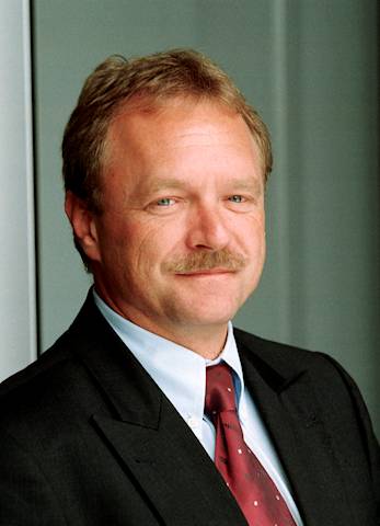 Ein Mann mit blondem Haar trägt einen dunkelgrauen Anzug und eine rot gepunktete Krawatte. Er steht vor einem neutralen Hintergrund. Der Kragen seines weißen Hemdes ist sichtbar.
