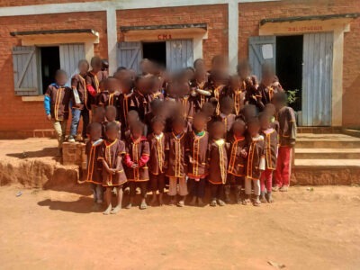 Eine große Gruppe von Kindern steht vor einem Gebäude und posiert für ein Foto. Sie tragen einheitliche braune Schuluniformen mit orangefarbenen Verzierungen. Das Gebäude im Hintergrund hat mehrere Fenster und eine einfache Architektur.