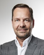 Ein Mann trägt ein weißes Hemd und ein graues Sakko. Der Hintergrund ist hell und neutral, wodurch das Outfit hervorgehoben wird. Der Mann hat kurzes, dunkles Haar.