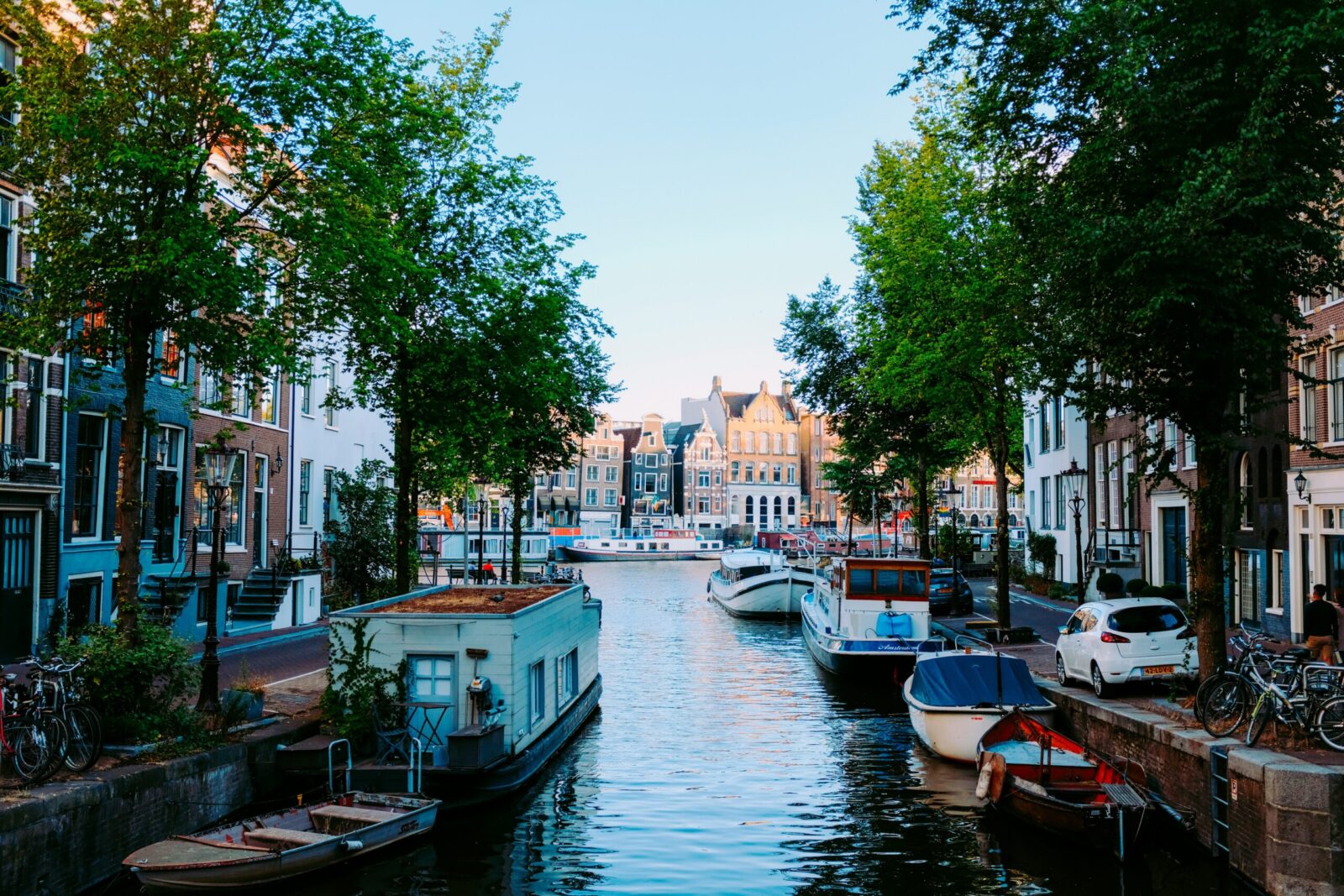 Bild eines malerischen Kanals in Amsterdam, gesäumt von Bäumen und traditionellen Häusern. Boote sind entlang des Wassers geparkt, und Fahrräder stehen am Ufer. Es herrscht eine ruhige und entspannte Atmosphäre bei klarem Himmel.