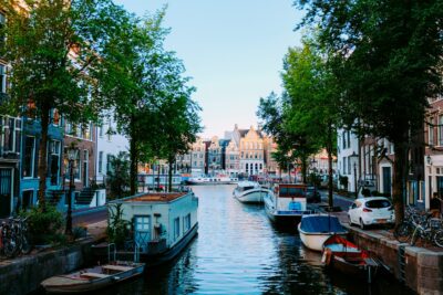 Bild eines malerischen Kanals in Amsterdam, gesäumt von Bäumen und traditionellen Häusern. Boote sind entlang des Wassers geparkt, und Fahrräder stehen am Ufer. Es herrscht eine ruhige und entspannte Atmosphäre bei klarem Himmel.