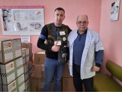 Zwei Personen stehen in einem Raum voller Kartons und lächeln in die Kamera. Die Wände des Raumes sind rosa gestrichen und ein Plakat hängt an der Wand. Einer der Personen trägt eine weiße Arbeitskittel, während die andere einen Pullover mit verschiedenen Abzeichen trägt.