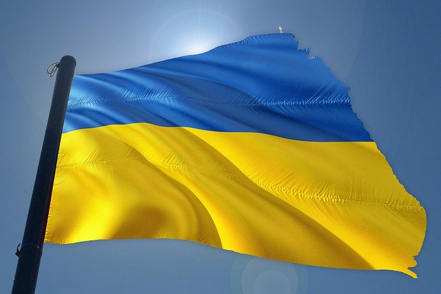 Die Flagge der Ukraine weht im Wind vor einem klaren blauen Himmel. Die Flagge besteht aus zwei horizontalen Streifen, oben blau und unten gelb. Im Hintergrund scheint die Sonne, die die Farben der Flagge lebendig erscheinen lässt.