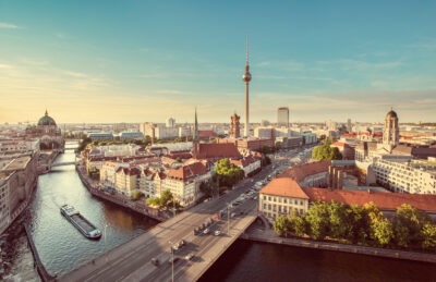 Ein Blick über Berlin zeigt den Fernsehturm und andere markante Gebäude. Die Stadt erstreckt sich bis zum Horizont unter einem blauen Himmel. Autos fahren über eine große Brücke, die über einen Fluss führt.