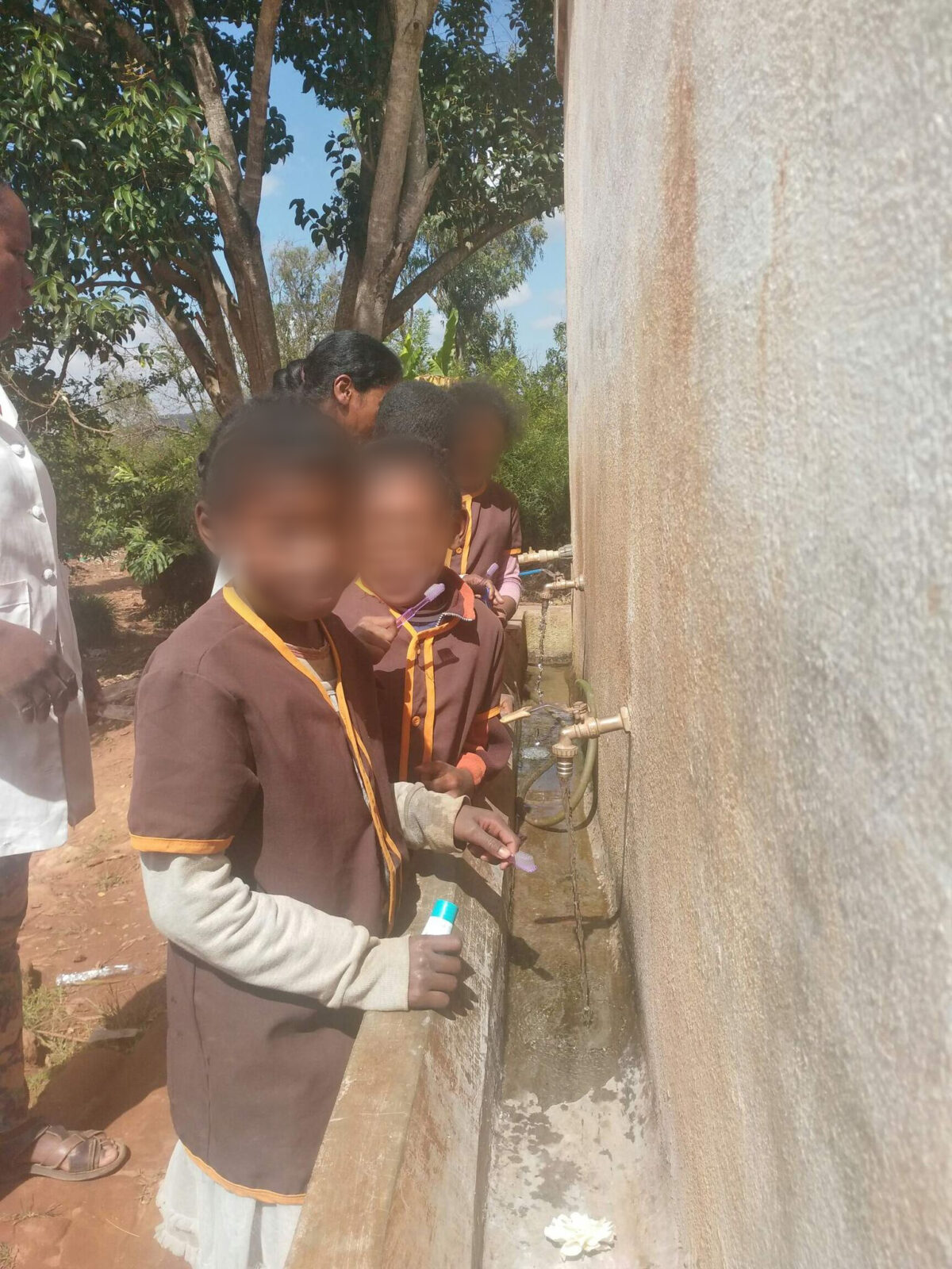 Zwei Kinder stehen an einem Wasserhahn und füllen ihre Flaschen. Sie tragen braune Uniformen mit orangefarbenen Akzenten. Die Umgebung wirkt ländlich und einfach.