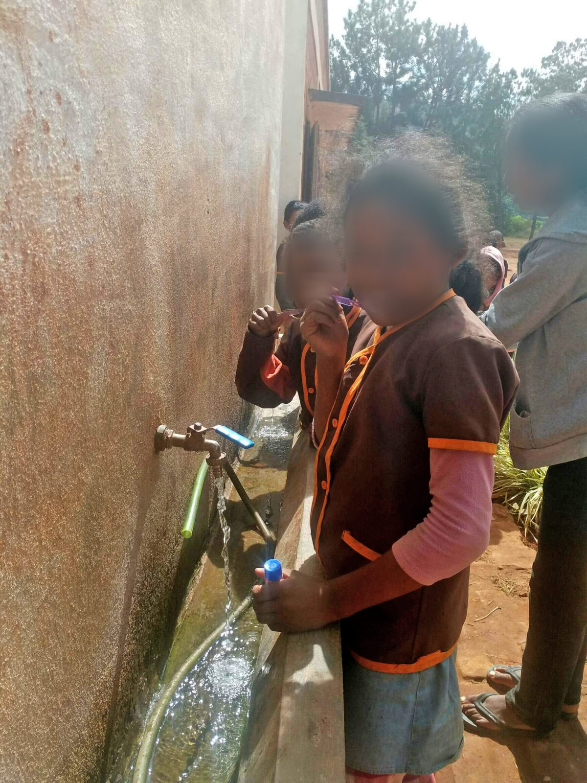 Eine Gruppe von Kindern steht an einer Wasserstelle und füllt ihre Flaschen. Sie tragen ähnliche braune Uniformen und scheinen miteinander zu sprechen. Die Wand neben ihnen ist aus Beton und zeigt leichte Abnutzungserscheinungen.