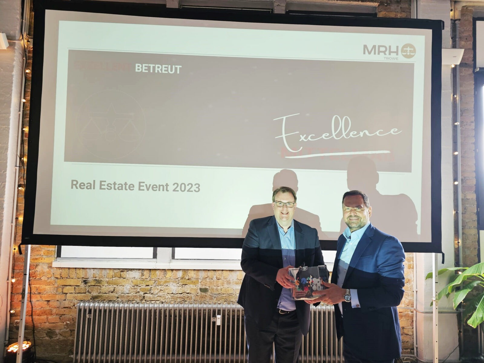 Zwei Personen stehen vor einer Leinwand, die die Aufschrift "Real Estate Event 2023" und "Excellence" zeigt. Die Veranstaltung scheint von MRH Trowe organisiert zu sein. Die Personen halten eine Auszeichnung.