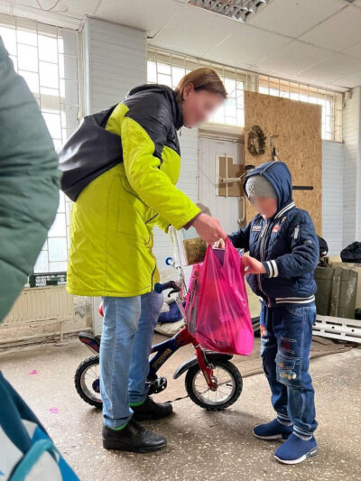 Eine Person in einer gelben Jacke überreicht einem Kind eine pinke Tasche. Das Kind steht neben einem kleinen roten Fahrrad und trägt eine blaue Jacke und Jeans. Im Hintergrund sind Fenster und eine Holzwand zu sehen.