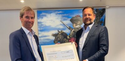 Zwei Männer in Anzügen halten einen großen Scheck über 3.200 Euro vor einem Gemälde, das einen Affen in historischer Kleidung auf einem Pferd zeigt. Der Scheck ist von der Charitybank GmbH und adressiert an die KiNiKi gAG.