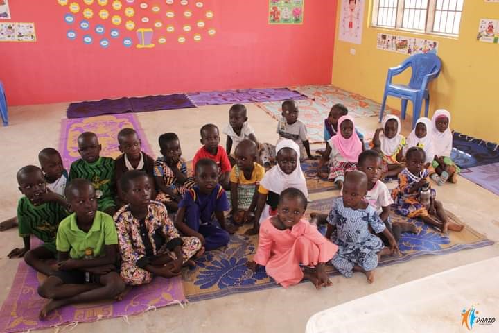 Kinder sitzen in einem Klassenzimmer auf bunten Matten, umgeben von farbenfrohen Wanddekorationen und Lernmaterialien.
