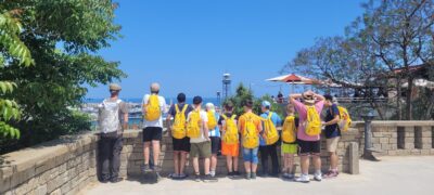 Eine Gruppe von Kindern steht auf einer Aussichtsplattform und blickt auf eine Stadtlandschaft mit einem Turm im Hintergrund. Die Kinder tragen gelbe Rucksäcke und sind sommerlich gekleidet. Die Szene ist bei klarem, sonnigem Wetter aufgenommen, und im Hintergrund sind grüne Pflanzen und ein Café mit Sonnenschirmen zu sehen.