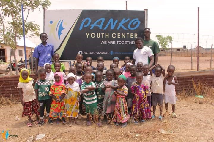 Eine Gruppe von Kindern steht vor einem Schild des Panko Youth Centers in einer trockenen Umgebung. Zwei Erwachsene stehen links und rechts neben den Kindern. Das Schild trägt die Aufschrift "Panko Youth Center - We are together" und zeigt Kontaktinformationen. Die Kinder tragen bunte Kleidung und scheinen Teil einer Gemeinschaft oder Veranstaltung zu sein.