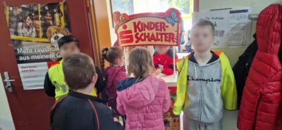 Eine Gruppe von Kindern steht vor einem Schalter mit der Aufschrift "Kinderschalter" in einer Bank. Im Hintergrund ist ein Plakat mit der Aufschrift "Mehr rausholen aus meinem Geld" und eine Tür zu sehen. An der Wand hängt ein weiteres Informationsblatt.