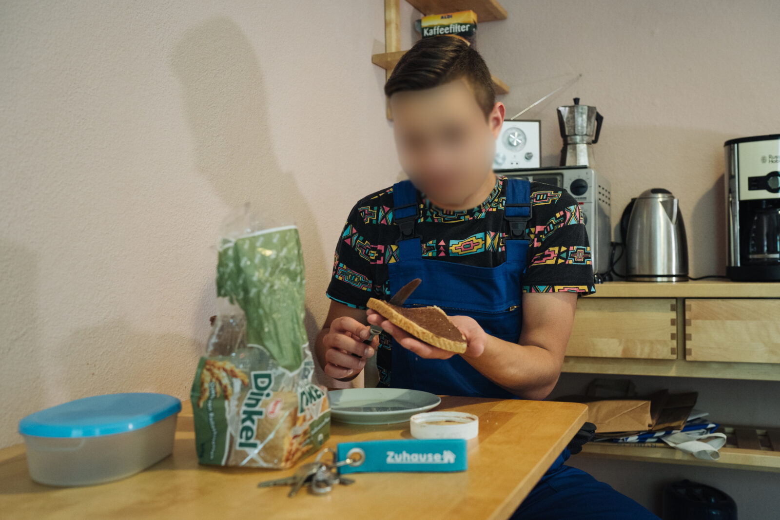 Eine Person in blauer Latzhose sitzt an einem Küchentisch und bereitet ein Brot zu. Auf dem Tisch befinden sich eine Packung Dinkelbrot, ein Teller, eine Brotdose, ein Schlüsselbund und eine Rolle Klebeband. Im Hintergrund sind Küchengeräte wie eine Kaffeemaschine und ein Wasserkocher zu sehen.