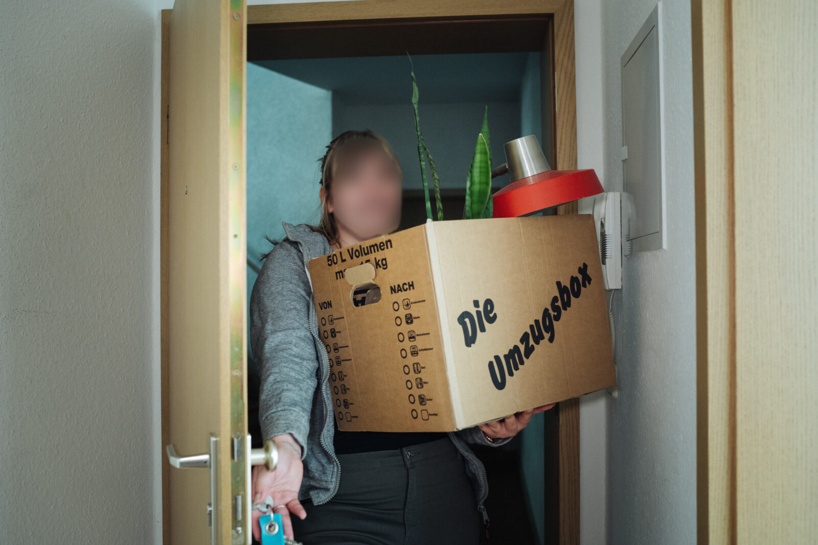 Person trägt einen großen Umzugskarton mit der Aufschrift "Die Umzugsbox" durch eine Tür, darin sind eine Pflanze und eine rote Lampe zu sehen.