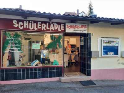 Außenansicht eines kleinen Ladens namens "Schülerlädle" mit einem Schaufenster, das Brötchentheke, Getränke, Süßigkeiten, Schreibwaren und vieles mehr anbietet. Durch die offene Tür sind Personen im Inneren des Ladens zu sehen.