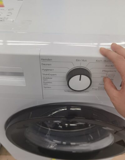 Nahaufnahme einer Waschmaschine mit einem Bedienfeld, auf dem verschiedene Waschprogramme wie Hemden, Daunen, Hygiene+, StainExpert, Outdoor, Dunkles, Spülen, Koch-/Buntwäsche, Eco 40-60, Pflegeleicht, Express, Feinwäsche und Schleudern/Abpumpen zu sehen sind. Eine Hand dreht den Programmwähler.