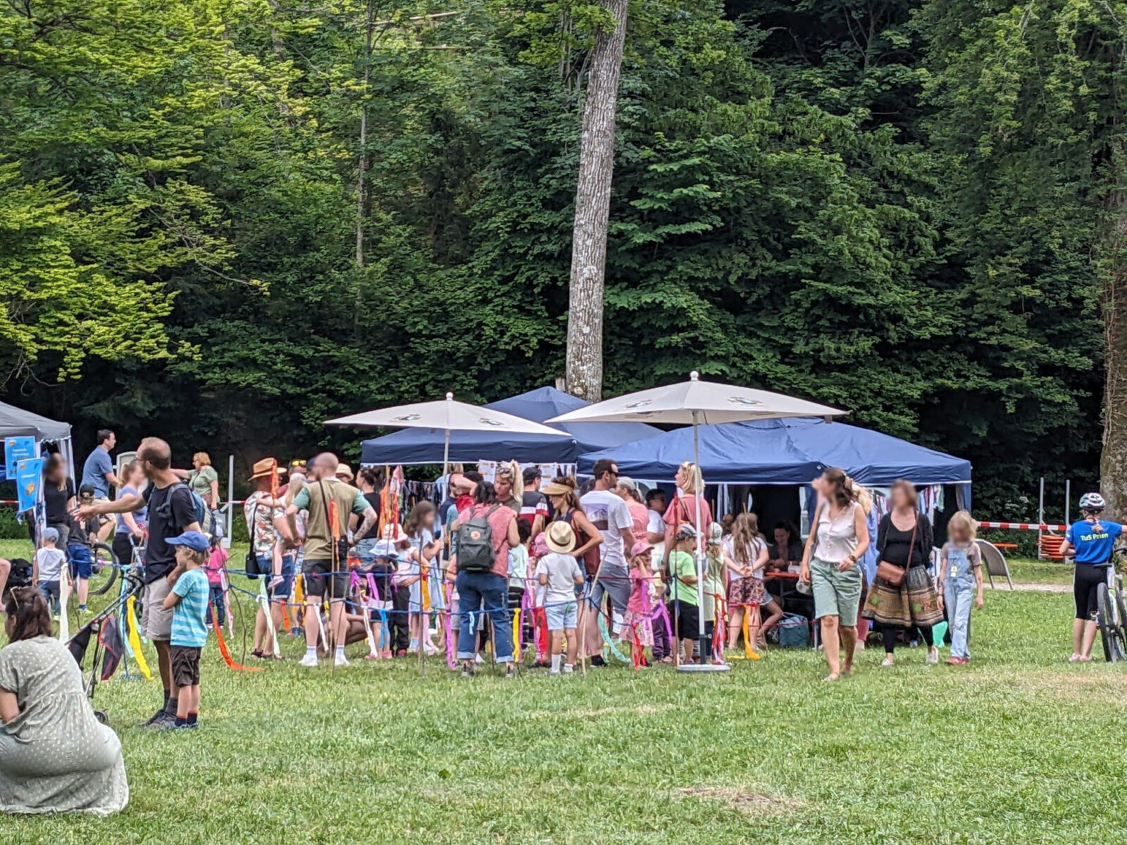 Eine Gruppe von Menschen, darunter viele Familien mit Kindern, versammelt sich bei einem Outdoor-Event auf einer grünen Wiese. Es gibt mehrere blaue Pavillons und Sonnenschirme. Der Hintergrund ist von üppigen grünen Bäumen umgeben.