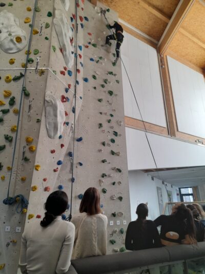 Eine Person klettert eine hohe Indoor-Kletterwand mit bunten Griffen hinauf, während vier Zuschauer am Boden sitzen und zuschauen.