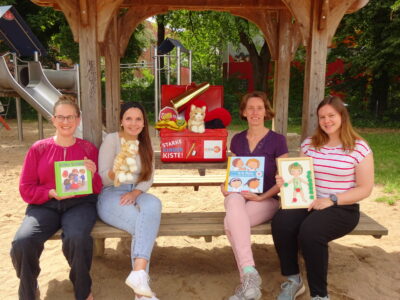 Vier Frauen sitzen auf einer Holzbank unter einem Pavillon auf einem Spielplatz und halten Kinderbücher in den Händen. Hinter ihnen steht eine rote Box mit der Aufschrift "Starke Kinder Kiste!" und verschiedenen Spielzeugen. Im Hintergrund sind Bäume und ein Klettergerüst zu sehen.