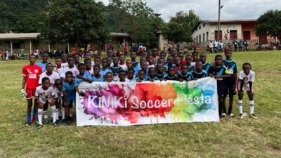 Eine große Gruppe von Kindern in Fußballtrikots steht auf einem Spielfeld und hält ein buntes Banner mit der Aufschrift "KiNiKi Soccer Fiesta" vor sich. Im Hintergrund sind Zuschauer und Gebäude zu sehen.