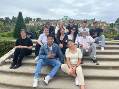 Gruppe von Menschen sitzt auf Steintreppen im Freien vor einem historischen Gebäude mit grünen Gärten im Hintergrund.