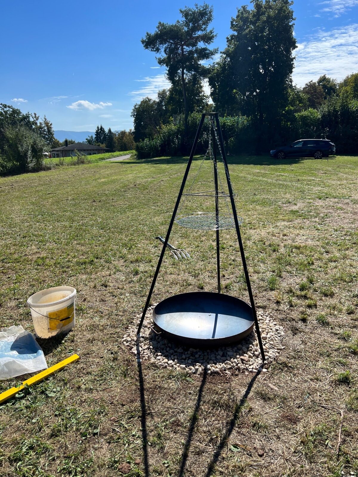 Ein dreibeiniger Grillständer mit einer großen Pfanne und einem Grillrost, aufgestellt auf einer Wiese unter klarem blauem Himmel. Im Hintergrund sind Bäume, ein geparktes Auto und ein Haus zu sehen. Links neben dem Grillständer stehen ein weißer Eimer und verschiedene Werkzeuge auf dem Boden.