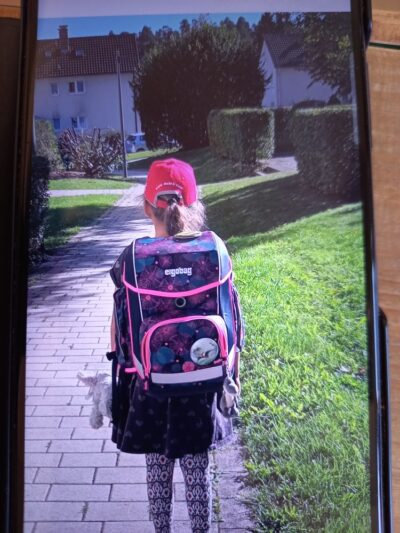 Ein Mädchen mit rotem Cappy und großem Schulrucksack geht auf einem gepflasterten Weg entlang, umgeben von Grünflächen und Wohnhäusern im Hintergrund.