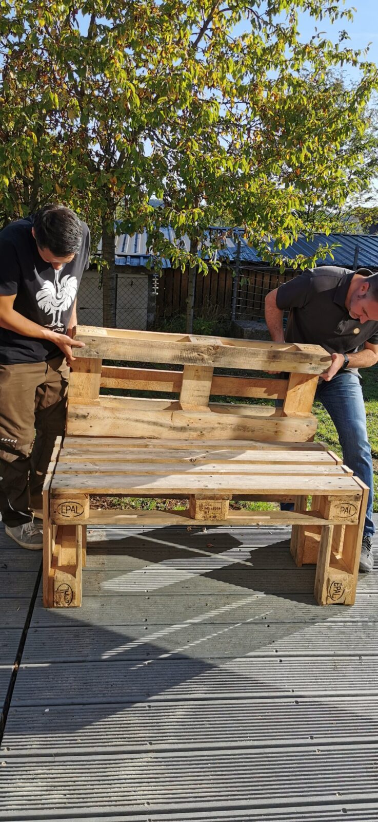 Zwei Männer bauen eine Bank aus Holzpaletten auf einer Terrasse zusammen. Im Hintergrund sind Bäume und ein Gebäude zu sehen.