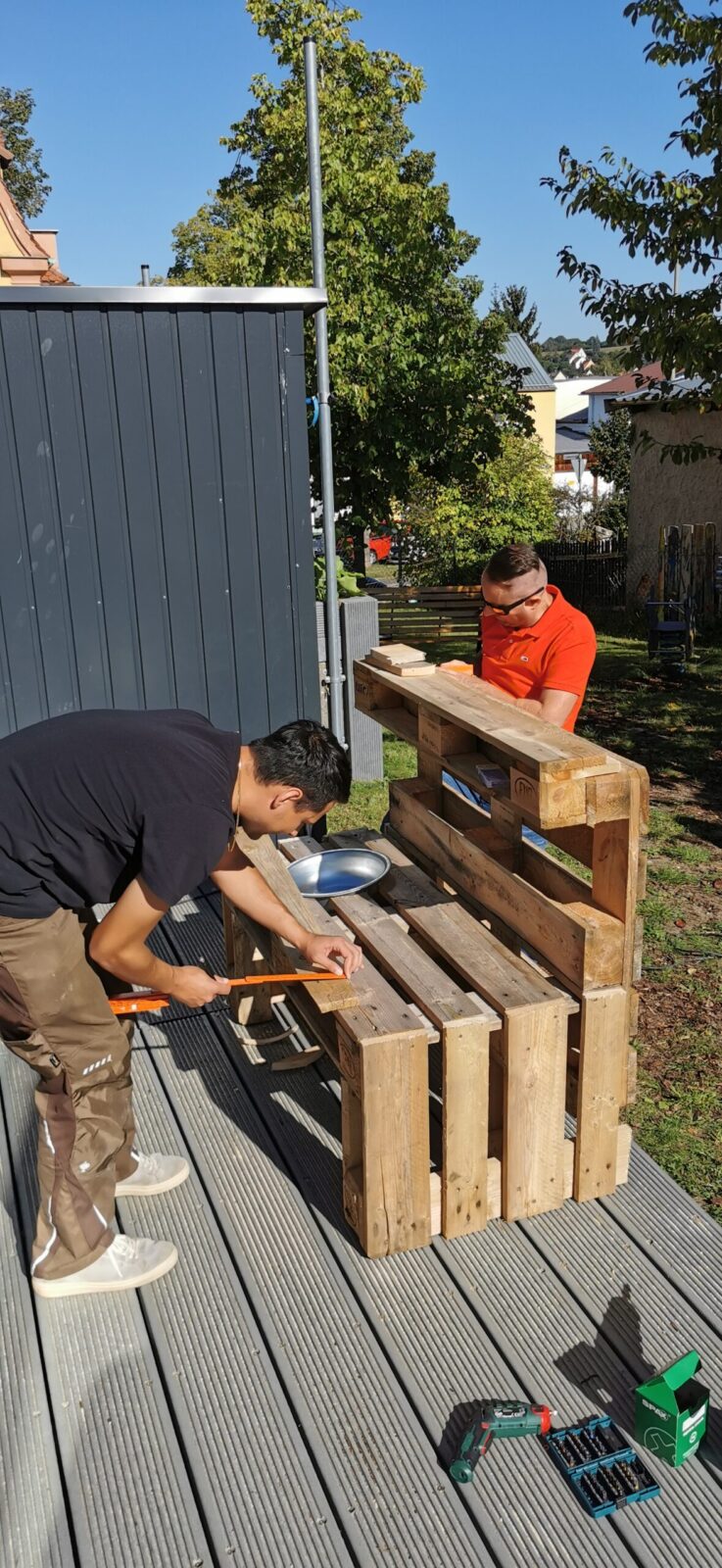 Zwei Männer bauen im Freien eine Bank aus Holzpaletten zusammen. Einer trägt ein schwarzes T-Shirt und arbeitet mit einem Hammer, während der andere ein orangefarbenes T-Shirt trägt und die Konstruktion stabilisiert. Im Hintergrund sind Bäume und Gebäude zu sehen. Werkzeuge liegen auf dem Boden.