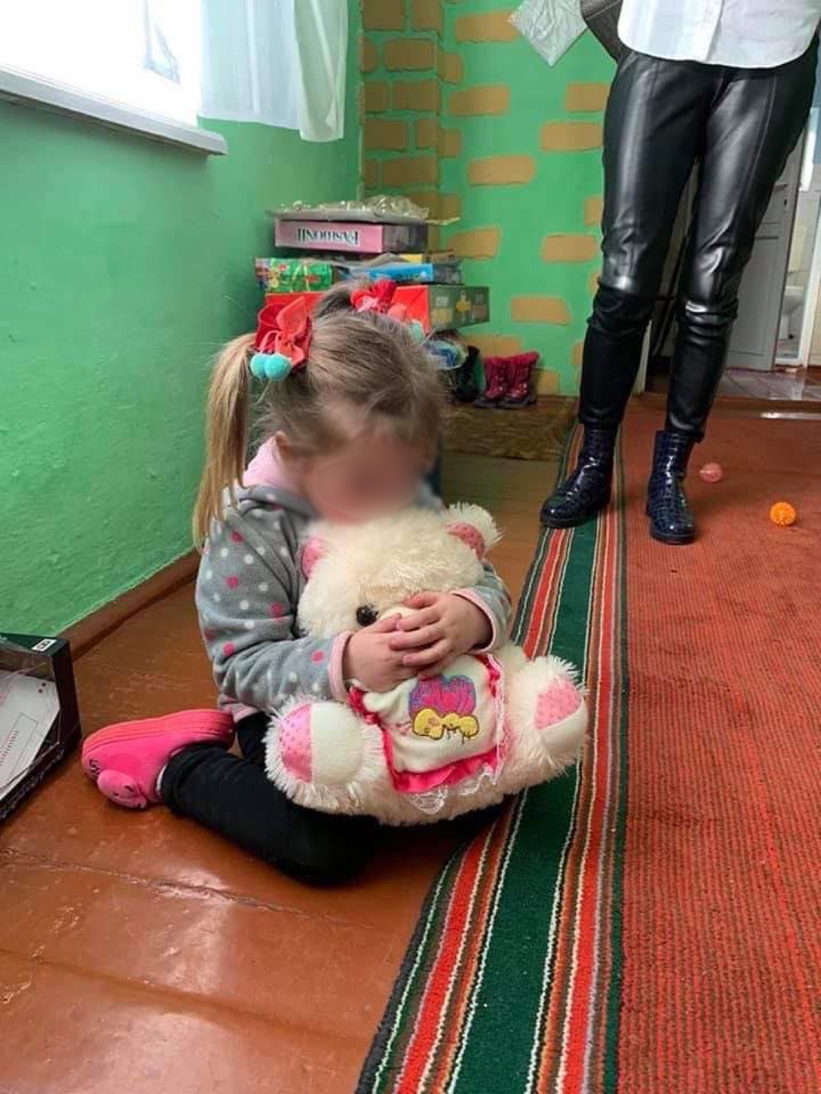 Ein kleines Mädchen mit roten Schleifen im Haar sitzt auf einem Teppich und umarmt einen weißen Plüschbären. Im Hintergrund stehen eine Person in schwarzen Lederhosen und verschiedene Spielsachen.