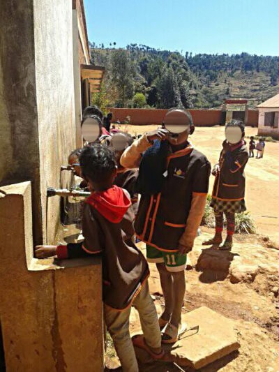 Kinder in Schuluniformen trinken Wasser aus einem öffentlichen Wasserhahn an einem sonnigen Tag in einer ländlichen Gegend. Im Hintergrund sind weitere Kinder und Gebäude zu sehen.