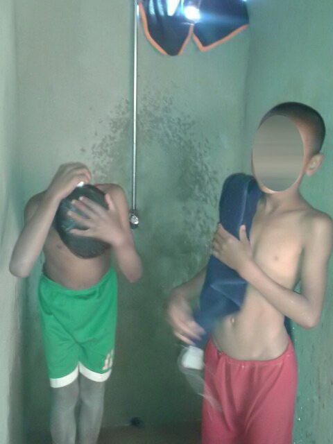 Zwei Jungen stehen in einer Dusche, wobei einer seinen Kopf mit Wasser bedeckt und der andere ein Handtuch hält.
