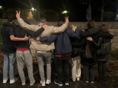 Eine Gruppe von sieben Personen steht eng umarmt und mit dem Rücken zur Kamera, einige zeigen das "Peace" mit ihren Händen oder Daumen hoch. Sie stehen draußen bei Nacht mit einer Steinmauer im Hintergrund.