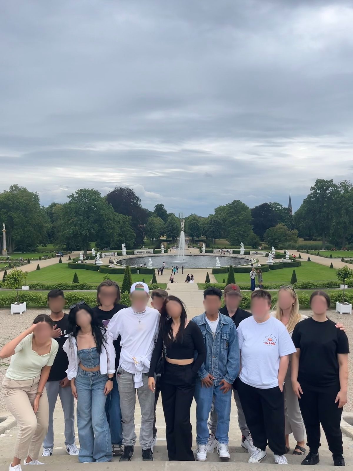 Eine Gruppe von Menschen steht in einem Park vor einem großen Brunnen und einer gepflegten Gartenanlage mit Statuen im Hintergrund.