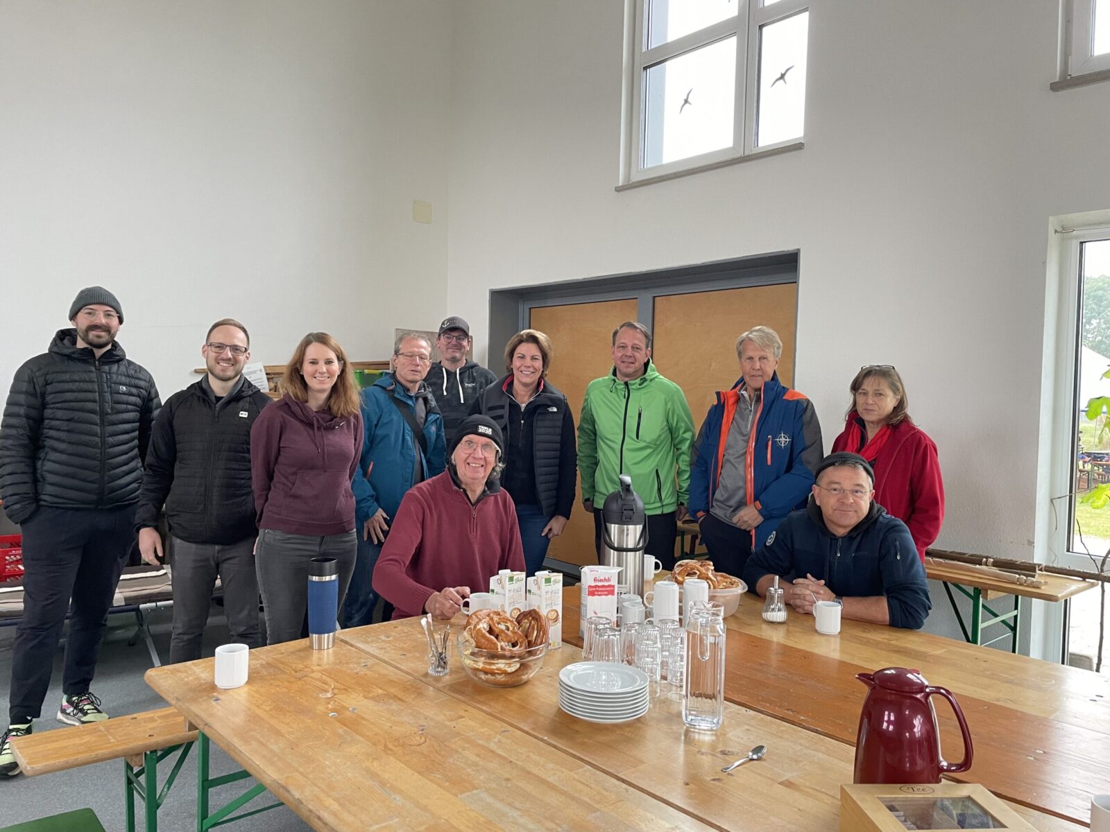 Eine Gruppe von Menschen steht in einem Raum, einige sitzen an einem Holztisch, der mit Frühstücksartikeln wie Brezeln, Getränkekartons, einer Kaffeekanne und Geschirr gedeckt ist. Im Hintergrund sind Fenster und eine Tür zu sehen.