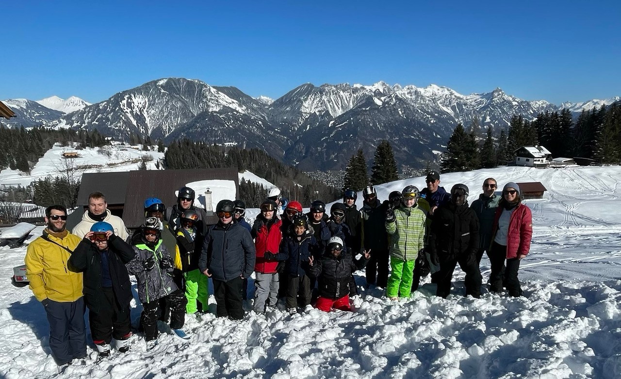 Eine Gruppe von Menschen in Winterkleidung steht auf einer schneebedeckten Fläche vor einer malerischen Berglandschaft mit klar blauem Himmel. Im Hintergrund sind Hütten und schneebedeckte Bäume zu sehen.