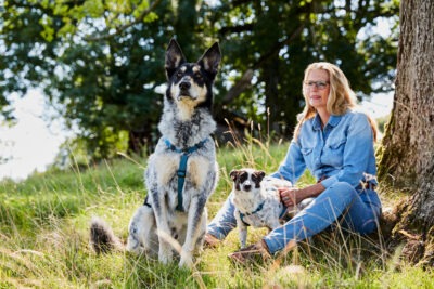Eine Frau in Jeanskleidung sitzt im Gras neben einem Baum, während zwei Hunde, einer groß und einer klein, in der Nähe sitzen. Die Szene spielt sich in einer natürlichen Umgebung mit grünem Gras und Bäumen im Hintergrund ab.