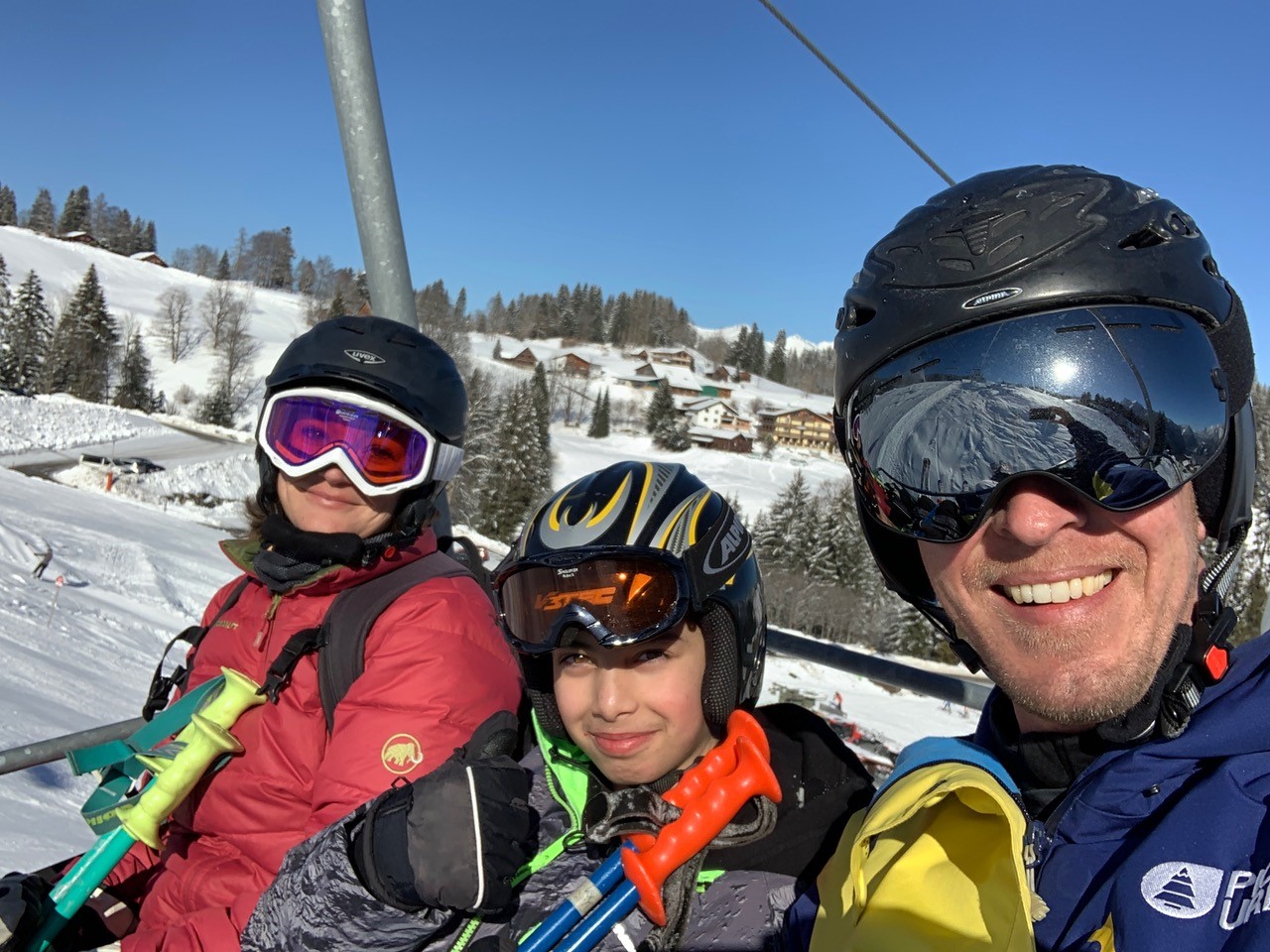 Drei Personen in Skiausrüstung sitzen auf einem Sessellift, umgeben von schneebedeckten Bergen und Bäumen.