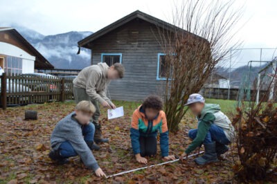 Kinder führen eine Outdoor-Aktivität durch, bei der sie auf dem Boden knien und ein Maßband verwenden, während ein weiteres Kind ihnen Anweisungen gibt und ein Blatt Papier hält. Im Hintergrund ist ein Holzhaus und eine Berglandschaft zu sehen.