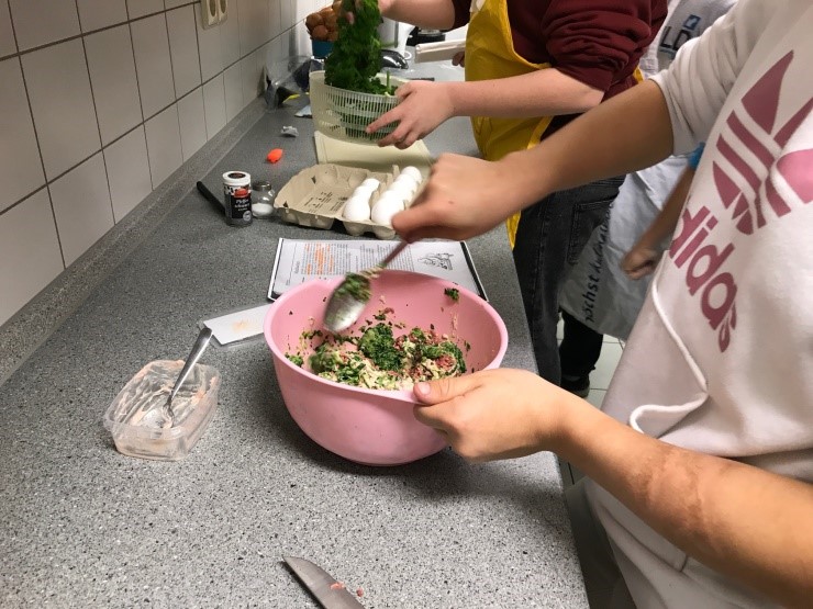 Personen bereiten in einer Küche eine Mahlzeit zu. Eine Person rührt in einer rosa Schüssel eine Mischung aus Kräutern und anderen Zutaten, während eine andere Person frische Kräuter aus einem Sieb nimmt. Auf der Arbeitsfläche liegen Eier, ein Rezept und verschiedene Kochutensilien.